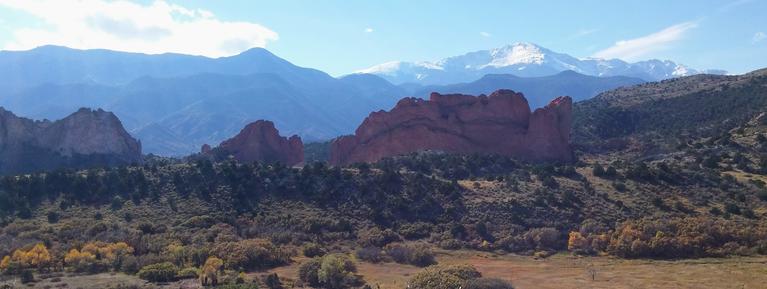 View of Pikes Peak from the North and Garden of the Gods between. The sky is blue with some clouds and the mountains are a cornflower blue against the red rocks of garden of the Gods.